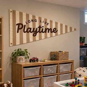 Könnte beinhalten: Ein großer dreieckiger Wandwimpel mit vertikalen beigen und weißen Streifen und dem Text "LONG LIVE Playtime" in dunkelbrauner Schrift. Der Wimpel hat Metallösen an den Ecken zur Befestigung an einer Wand über einem Spielzeugregal.