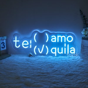 May include: A blue neon sign with the words "( )amo (✔) quila tei (V)" and the word "Purok" in a smaller font below. The sign is on a white fluffy surface.
