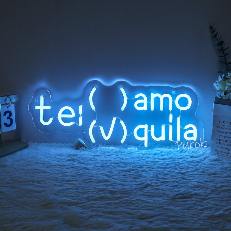 以下が含まれることがあります： 「( )amo (✔) quila tei (V)」という文字と、その下に小さく「Purok」という文字が書かれた青いネオンサイン。白いふわふわした表面の上に置かれている。