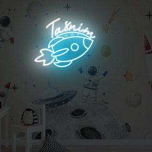 May include: A blue neon sign with a rocket ship and the name Tasnim in white neon lettering. The sign is on a white wall with a space-themed design.