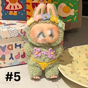 May include: A plush doll in a bunny costume with a green fuzzy body, a painted face, and flower accents. The doll wears a yellow crocheted bikini bottom and a floral headband. The number "#5" is visible in the lower left corner.