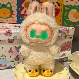 May include: A plush toy doll in a bunny costume with a smiling face and green glasses. The doll is wearing a fluffy cream-colored outfit with matching bunny ears. The doll's feet are yellow with black dots. The doll is on a yellow plate.