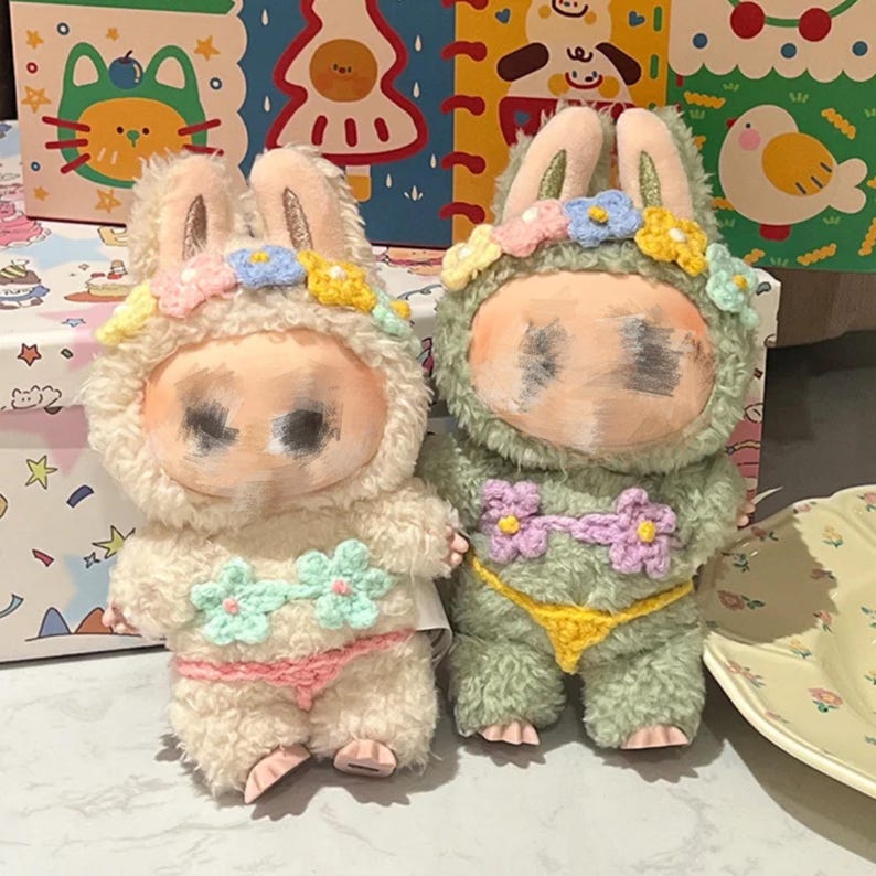 May include: Two plush bunny dolls, one cream and one green, each wearing a floral crown and crochet bikini. The cream bunny has a pink bikini, while the green bunny has a yellow bikini. The dolls have painted faces and are standing upright.