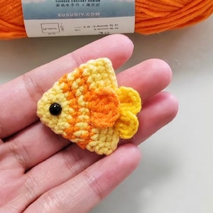 May include: A handmade crochet fish in yellow and orange, with a black eye. The fish is held in a hand, with a ball of orange yarn in the background. The fish is approximately 1.25 inches long.