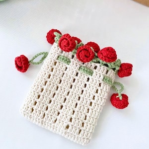 以下が含まれることがあります： 赤いバラのトリムが付いた白いクロシェの巾着袋。バッグは繊細なオープンウィーブのデザインで、小さな赤いバラと緑の葉で飾られています。