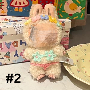 May include: A plush bunny toy with a cream-colored body, wearing a pink bikini and a flower crown. The bunny has a partially obscured face and is posed in front of a patterned box. The number "#2" is visible.