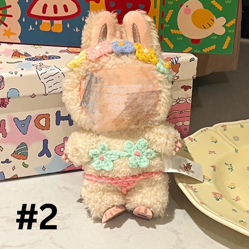 May include: A plush bunny toy with a cream-colored body, wearing a pink bikini and a flower crown. The bunny has a partially obscured face and is posed in front of a patterned box. The number "#2" is visible.