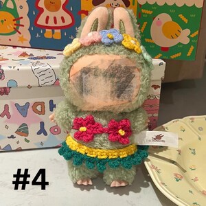 May include: A plush toy in the shape of a green bunny wearing a floral crown and a crochet outfit with red flowers. The number "#4" is displayed in black at the bottom left.