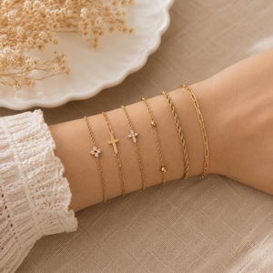 May include: A set of six gold-toned bracelets featuring various designs including a four-stone floral charm, a simple cross, a small crystal cross, a delicate chain with two beads, a twisted rope chain, and a classic oval link chain.