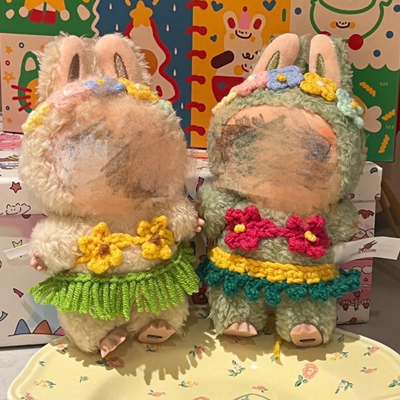 May include: Two plush bunny dolls with floral headbands and crochet skirts. One bunny is cream-colored with a green skirt, the other is green with a red skirt. Both dolls have a yellow floral necklace.