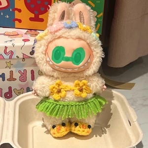 May include: A plush toy doll with a bunny-eared hood, floral accents, and a green-tinted face. The doll wears a yellow flower top and a green skirt. Its feet are yellow with black dots. The doll is in a white container.