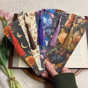 May include: A collection of decorative bookmarks with colorful tassels. Each bookmark features a unique design, including floral patterns, butterflies, and coffee cup illustrations. The bookmarks are held over an open book.