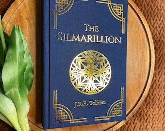 May include: A navy blue hardcover book titled "The Silmarillion" by J.R.R. Tolkien. The book features gold foil lettering and decorative elements. A gold circular design is centered on the cover. The book is displayed on a wooden tray.