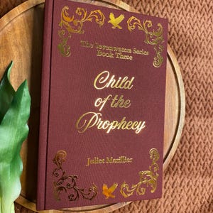 May include: A hardcover book titled Child of the Prophecy by Juliet Marillier, which is book three of The Sevenwaters Series. The book has a deep maroon fabric cover with gold foil lettering and ornate filigree designs featuring small birds.