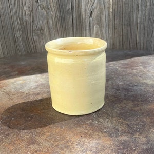 May include: A pale yellow ceramic jar with a rounded bottom and a slightly flared top. The jar is sitting on a rusty metal surface.