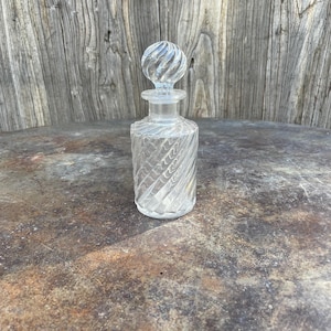 May include: A clear glass perfume bottle with a spiral design. The bottle has a round, textured stopper. The bottle is approximately 10 cm tall.