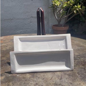 May include: Two white rectangular ceramic trays stacked. The trays have a slightly curved design and are on a textured surface. A dark wooden object stands behind the trays, and a potted plant is in the background.