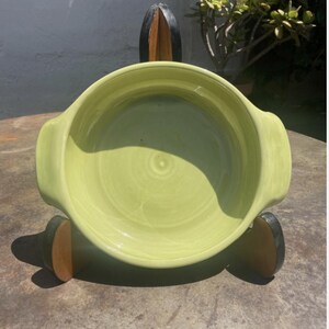 May include: A round, light green ceramic bowl with two integrated side handles, displayed upright on a dark-colored, three-pronged wooden stand. The bowl features a smooth, glossy finish with subtle, circular brushstroke patterns visible on the interior surface.