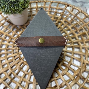 May include: A gray, pyramid-shaped box with an etched design and a brown leather strap secured by a gold-colored button. The box is resting on a woven, circular placemat, with a small potted plant in the background.
