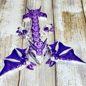 May include: A purple and white 3D printed dragon figurine with wings, a head, and a segmented body. The dragon is made of plastic and has a heart-shaped pattern on its body.