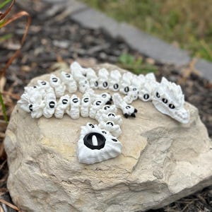 May include: White ceramic dragon figurine with black eyes. The dragon is curled up in a sleeping position. The figurine is on a rock.