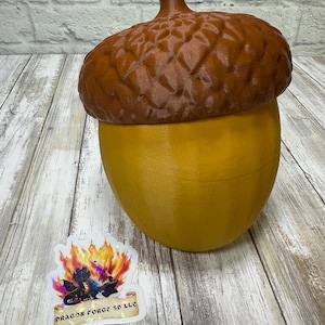May include: A large, decorative acorn-shaped container. The top is a textured brown, and the base is a smooth, golden yellow. The acorn has a stem and is sitting on a wooden surface. A sticker with the text "DRAGON FORGE 3D LLC" is visible.