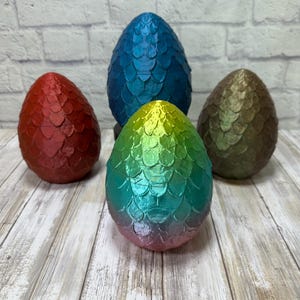 Op de afbeelding: Vier decoratieve draken eieren in verschillende kleuren: rood, blauw, groen en brons. Elk ei heeft een textuur, schubachtig patroon. De eieren zijn gerangschikt op een lichtgekleurd houten oppervlak, met een witte bakstenen achtergrond.