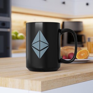 May include: Black ceramic coffee mug with a white Ethereum logo. The mug is sitting on a wooden surface.
