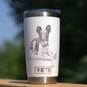 May include: A white stainless steel travel mug featuring a black and white etched illustration of a dog. The mug has a black lid and a silver base. The brand name "YETI" is printed on the front.