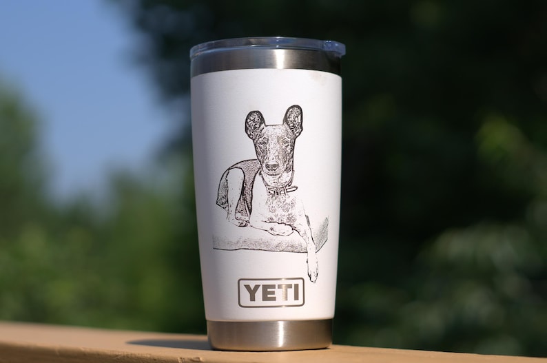 May include: A white stainless steel travel mug featuring a black and white etched illustration of a dog. The mug has a black lid and a silver base. The brand name "YETI" is printed on the front.