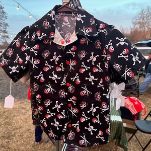 Può includere: Una camicia nera a maniche corte con bottoni e un motivo di teschi e ossa incrociate bianche. La camicia ha bottoni rossi e un colletto bianco.