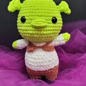 May include: Green crocheted plush toy with a white and brown outfit. The toy has large black eyes and a small, pointed nose.