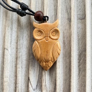 Op de afbeelding: Een lichtbruine houten uilenhanger met uitgesneden vleugeldetails en grote ronde ogen. De hanger hangt aan een zwart koord met twee verstelbare schuifknopen en een enkele donkerbruine houten kraal boven de uilenfiguur.