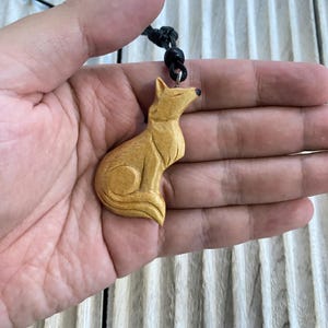 Puede incluir: Un collar con colgante de madera con un zorro tallado. El zorro marrón claro está suspendido de un cordón negro. El colgante se sostiene en una mano sobre un fondo de madera clara. El diseño del zorro es detallado.