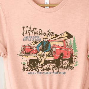 May include: Peach-colored t-shirt featuring a vintage-style illustration of a man sitting in the back of a red pickup truck. The text on the shirt reads: 'If I Had Two Dozen Roses and an Older Bottle of Wine, If I Really Coulda Hung The Moon, Would You Change Your Mind?' The design is suitable for country, western, or rustic themes.