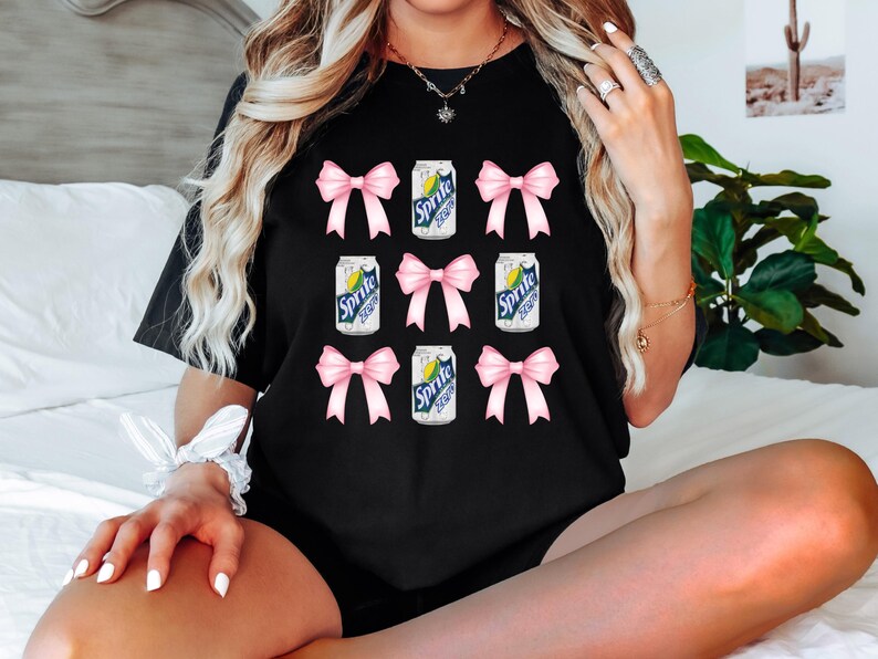 Op de afbeelding: Zwart T-shirt met een herhalend patroon van roze strikken en blikjes Sprite.