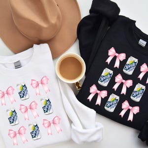Op de afbeelding: Twee witte en zwarte sweatshirts met een grafische print van roze strikken en blikjes Sprite. De tekst op de blikjes luidt "Sprite Lemon Lime".