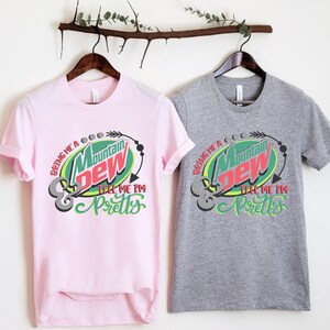Puede incluir: Dos camisetas con un logotipo de Mountain Dew y el texto "Bring me a Mountain Dew & tell me I'm pretty". La camiseta de la izquierda es rosa y la camiseta de la derecha es gris.