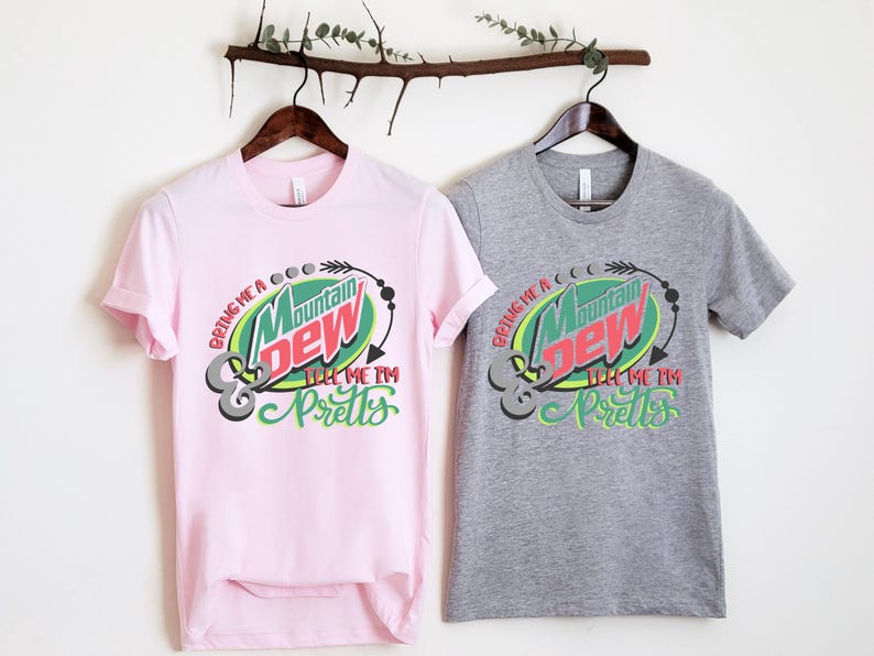 Puede incluir: Dos camisetas con un logotipo de Mountain Dew y el texto "Bring me a Mountain Dew & tell me I'm pretty". La camiseta de la izquierda es rosa y la camiseta de la derecha es gris.
