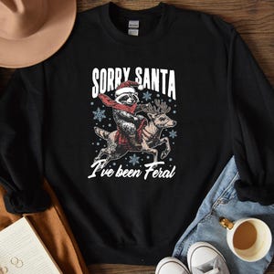 May include: Black sweatshirt with a white graphic of a raccoon riding a reindeer. The graphic says "Sorry Santa I've been Feral".