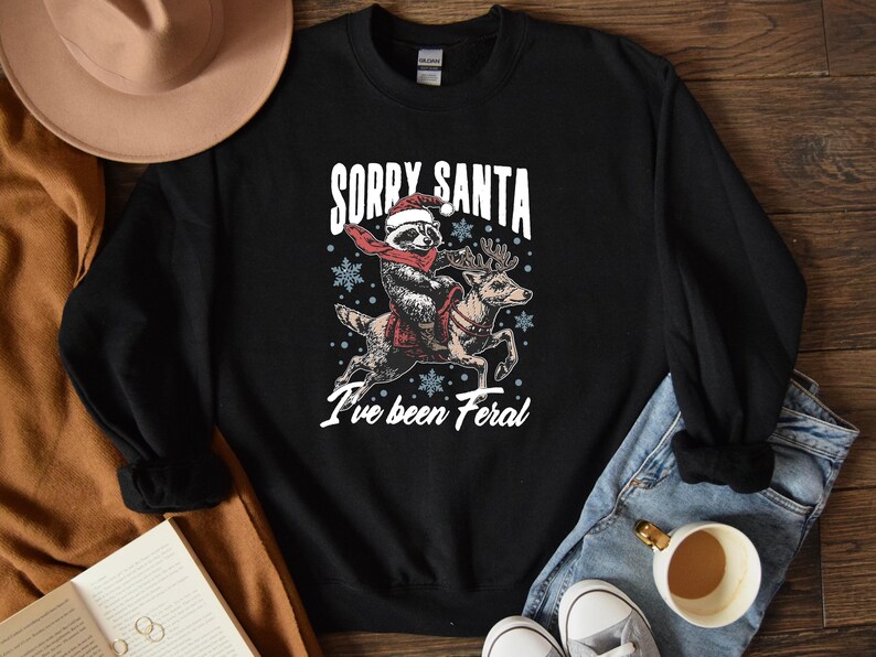 May include: Black sweatshirt with a white graphic of a raccoon riding a reindeer. The graphic says "Sorry Santa I've been Feral".