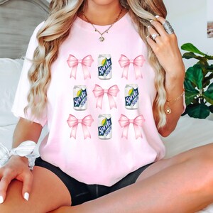 Op de afbeelding: Roze T-shirt met een grafische print van meerdere blikjes Sprite met roze strikken om elk blikje. De tekst op de blikjes luidt "Sprite".