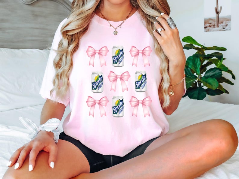 Op de afbeelding: Roze T-shirt met een grafische print van meerdere blikjes Sprite met roze strikken om elk blikje. De tekst op de blikjes luidt "Sprite".