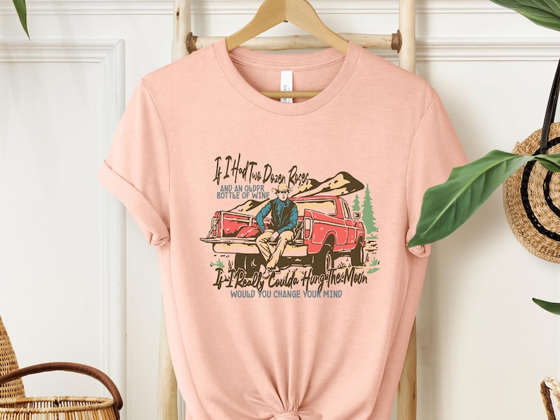 May include: Peach-colored t-shirt featuring a vintage-style illustration of a man sitting in the back of a red pickup truck. The text on the shirt reads: 'If I Had Two Dozen Roses and an Older Bottle of Wine, If I Really Coulda Hung The Moon, Would You Change Your Mind?' The design is suitable for country, western, or rustic themes.