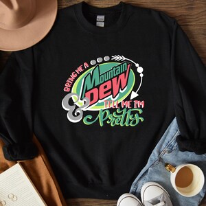 Puede incluir: Sudadera negra con un logotipo de Mountain Dew verde, rojo y blanco y el texto "Bring me a Mountain Dew & tell me I'm pretty".
