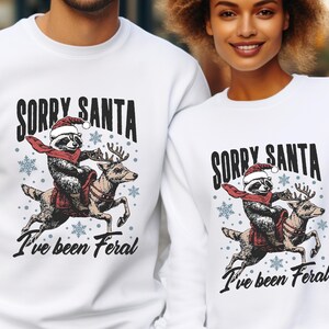 May include: White Christmas jumper featuring a graphic of a raccoon riding a reindeer. The text on the jumper reads "Sorry Santa I've been Feral".