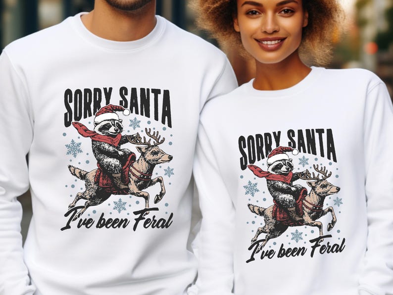 May include: White Christmas jumper featuring a graphic of a raccoon riding a reindeer. The text on the jumper reads "Sorry Santa I've been Feral".