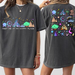 Op de afbeelding: Een donkergrijs T-shirt met een kleurrijke afbeelding van een kasteel en andere attractieparkiconen. De voorkant van het shirt zegt "Meet me at my happy place".