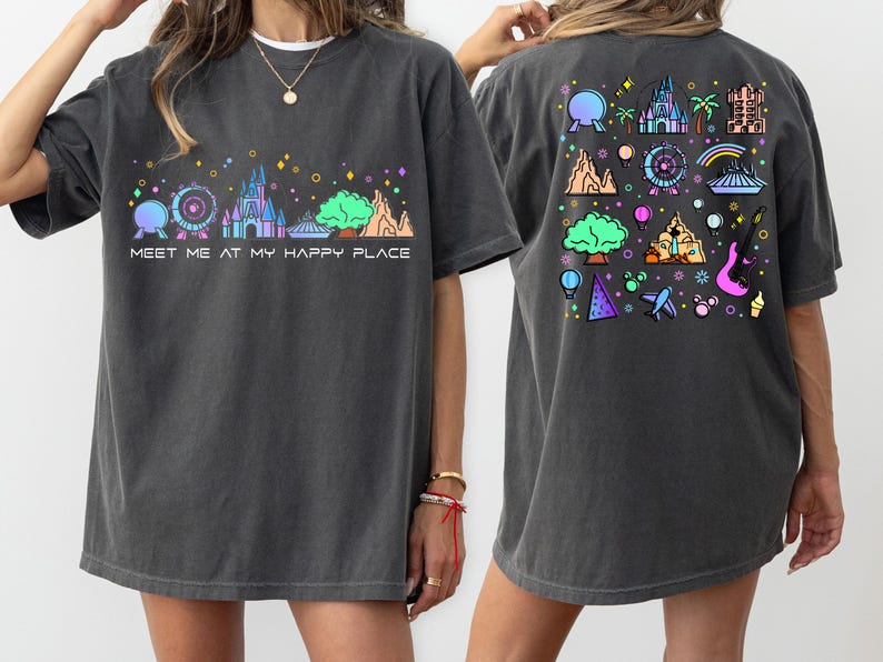 Op de afbeelding: Een donkergrijs T-shirt met een kleurrijke afbeelding van een kasteel en andere attractieparkiconen. De voorkant van het shirt zegt "Meet me at my happy place".