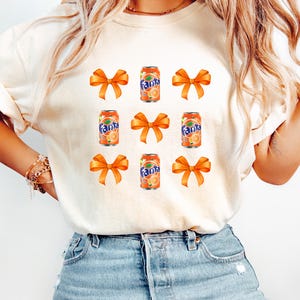 May include: A white t-shirt with a repeated pattern of orange soda cans and orange bows. The soda cans have the word "Fanta" printed on them.